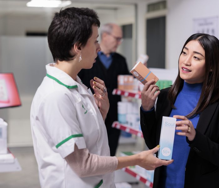 Pharmacist recommending woman sunblock cream in drugstore Pharmacist recommending young asian woman sunblock cream in drugstore. Customer holding skin care products packages, choosing between two sunscreen lotions and consulting with pharmacy assistant
