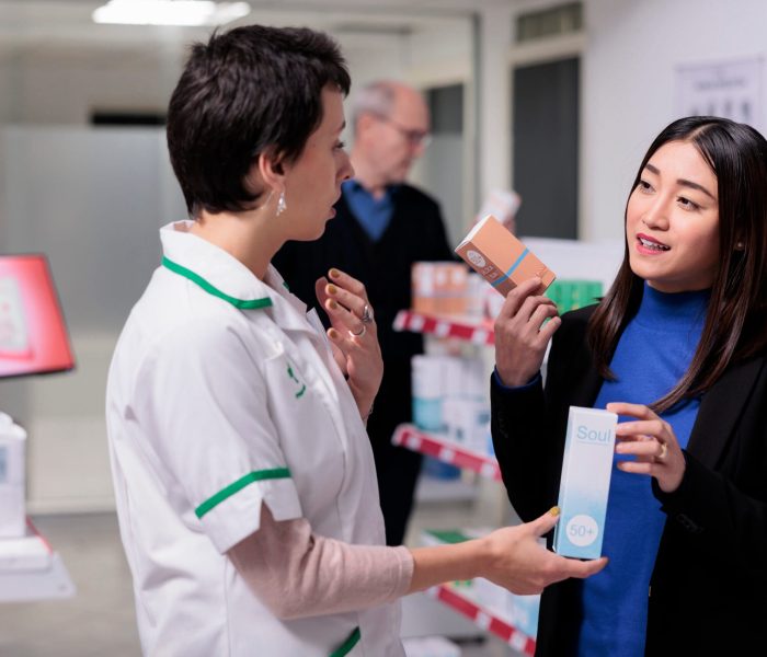 Pharmacist recommending young asian woman sunblock cream in drugstore. Customer holding skin care products packages, choosing between two sunscreen lotions and consulting with pharmacy assistant